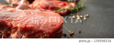Different cuts of beef steak are placed on a dark surface. Fresh herbs and spices are nearby, indicating preparation for cooking a meal in the kitchen, banner 139598316
