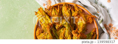 Golden-brown deep-fried zucchini flowers are arranged on a vibrant plate. The dish is set on a floral-patterned napkin, enhancing its appetizing appearance, banner Golden-brown deep-fried zucchini flowers are arranged on a vibrant plate. The dish is set on a floral-patterned napkin, enhancing its appetizing appearance, banner 139598317