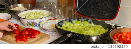 A woman cuts tomatoes on a wooden board surrounded by fresh vegetables while preparing squash caviar for winter canning in her kitchen, banner 139598329
