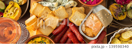 A vibrant charcuterie board showcases a variety of cheese, sausages, hummus, nuts, and sandwiches. Friends gather at a sunny outdoor picnic, enjoying delicious food together, banner A vibrant charcuterie board showcases a variety of cheese, sausages, hummus, nuts, and sandwiches. Friends gather at a sunny outdoor picnic, enjoying delicious food together, banner 139598340