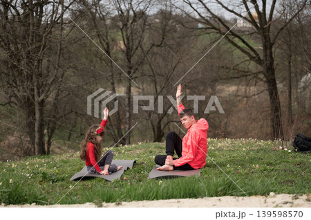 Two young individuals practicing yoga in a serene outdoor park during a cool spring afternoon surrounded by trees and nature 139598570