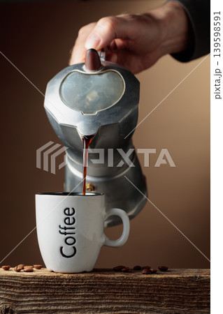 Black coffee is poured from a classic Italian aluminium moka pot into a coffee cup. Black coffee is poured from a classic Italian aluminium moka pot into a coffee cup. 139598591