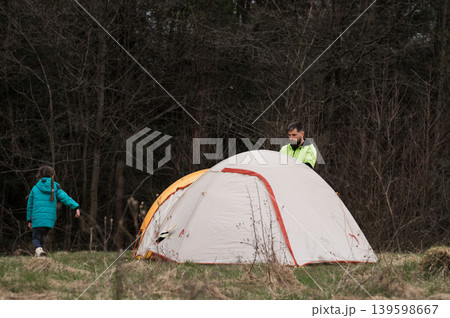 Family enjoys quiet camping experience in a serene forest setting as a child plays nearby and an adult sets up a cozy tent at dusk Family enjoys quiet camping experience in a serene forest setting as a child plays nearby and an adult sets up a cozy tent at dusk 139598667