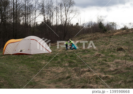 Camping adventure in the serene wilderness as hikers prepare their tent and gear on a grassy field surrounded by trees on a cloudy day Camping adventure in the serene wilderness as hikers prepare their tent and gear on a grassy field surrounded by trees on a cloudy day 139598668