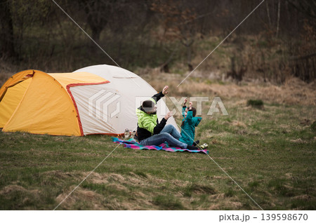 Joyful moments between a parent and child while camping in nature during a chilly afternoon, surrounded by lush greenery and a cozy tent Joyful moments between a parent and child while camping in nature during a chilly afternoon, surrounded by lush greenery and a cozy tent 139598670
