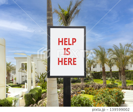 Help is here and support words symbol. Concept words Help is here on beautiful white sign. Beautiful hotel garden blue sky background. Business help is here and support concept. Copy space. 139599002
