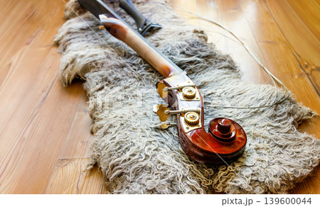 Broken contrabass neck joint with fingerboard, headstock with scroll, pegbox, tuning keys, nuts, pegs and brass plates lying on the wooden floor on felted wool locks rug.Copy space Broken contrabass neck joint with fingerboard, headstock with scroll, pegbox, tuning keys, nuts, pegs and brass plates lying on the wooden floor on felted wool locks rug.Copy space 139600044