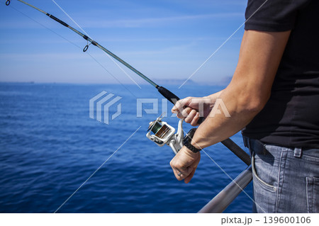 Close-up of man's hand holding fishing rod in the sea, man fishing. Fishing rod with a spinning reel in the hands of a fisherman. Close-up of man's hand holding fishing rod in the sea, man fishing. Fishing rod with a spinning reel in the hands of a fisherman. 139600106