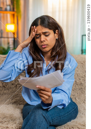 Indian woman in stress while looking at unpaid bills on home sofa with frustrated expression Indian woman in stress while looking at unpaid bills on home sofa with frustrated expression 139600419