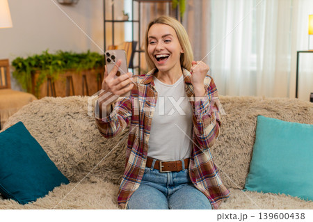 Adult woman smiling after reading message about salary raise and lottery win at home on smartphone Adult woman smiling after reading message about salary raise and lottery win at home on smartphone 139600438