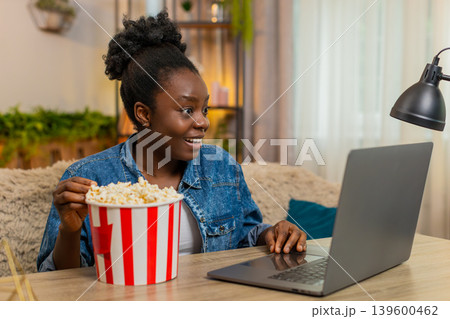 African American young woman watches comedy movie film on laptop with popcorn laughing at home room African American young woman watches comedy movie film on laptop with popcorn laughing at home room 139600462