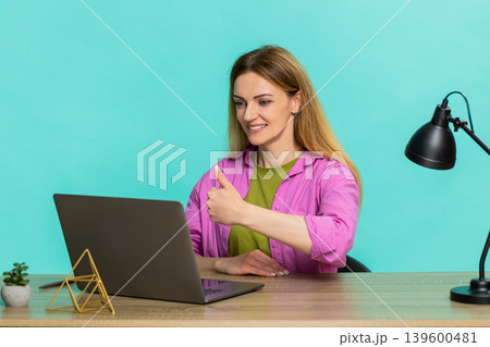 Adult woman sitting at table showing thumbs up to laptop webcam giving positive feedback with smile 139600481