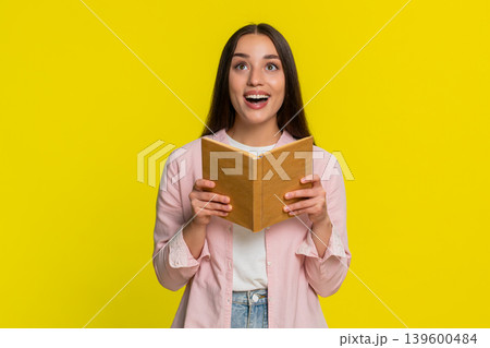 Young woman reading book with wide open eyes amazed expression intrigued thrilling surprising plot Young woman reading book with wide open eyes amazed expression intrigued thrilling surprising plot 139600484