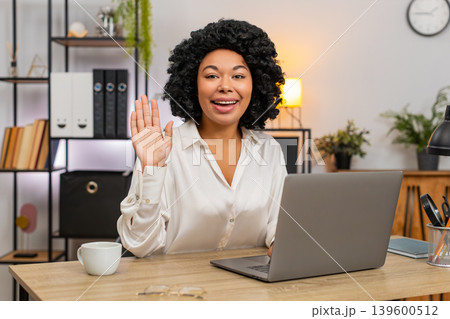 African American businesswoman waves hello toward laptop camera smiling friendly hi greeting gesture African American businesswoman waves hello toward laptop camera smiling friendly hi greeting gesture 139600512