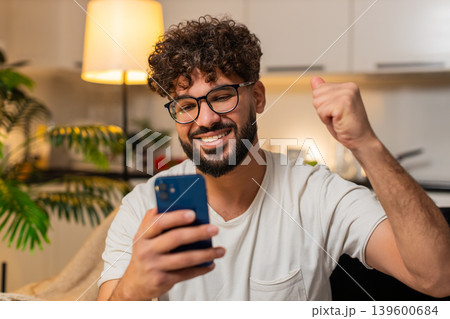 Arabian man celebrates online game jackpot win after good news alert on smartphone at night indoors 139600684
