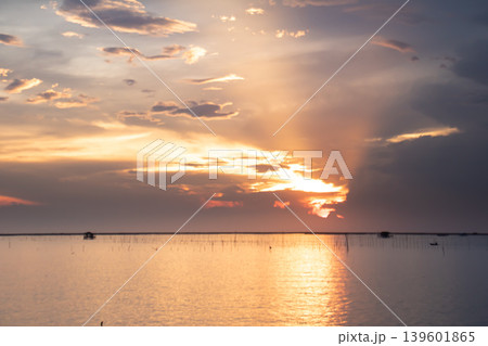 Gentle sunset light spreads across a wide lake, illuminating soft clouds and creating a warm golden reflection on calm water with distant structures along the horizon. Gentle sunset light spreads across a wide lake, illuminating soft clouds and creating a warm golden reflection on calm water with distant structures along the horizon. 139601865