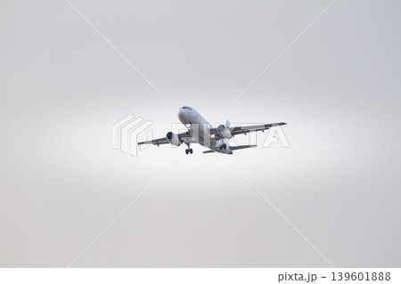 Modern twin engine passenger airplane flying toward landing approach against a soft cloudy sky at dusk with illuminated landing lights and extended landing gear. Modern twin engine passenger airplane flying toward landing approach against a soft cloudy sky at dusk with illuminated landing lights and extended landing gear. 139601888