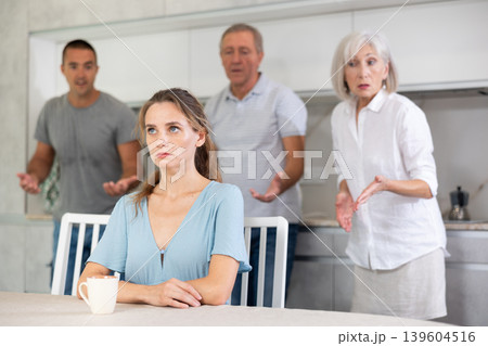 Domestic quarrel - offended young woman sits at table while parents and young husband yell at her 139604516