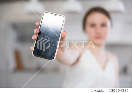 In home kitchen with white interior, smiling European girl in dress shows blank smartphone screen In home kitchen with white interior, smiling European girl in dress shows blank smartphone screen 139604583