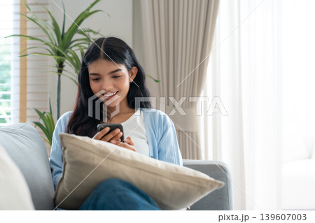 Smiling pretty young Asian woman sitting on cozy couch Smiling pretty young Asian woman sitting on cozy couch 139607003