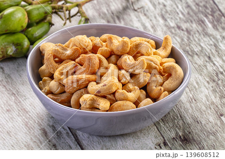 Roasted cashew nuts in a brown bowl with a cluster of raw green cashew fruit on a rustic wooden table. 139608512