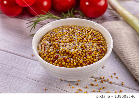 Whole grain mustard in white bowl, surrounded by fresh cherry tomatoes, rosemary, green onion on rustic wooden table 139608546