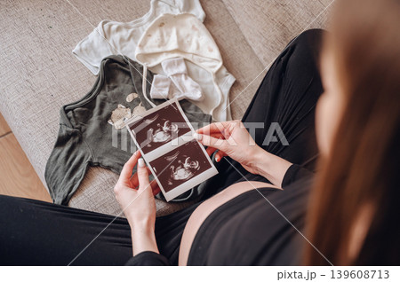 Close up of a pregnant woman holding ultrasound images of her baby next to tiny clothes on a sofa Close up of a pregnant woman holding ultrasound images of her baby next to tiny clothes on a sofa 139608713