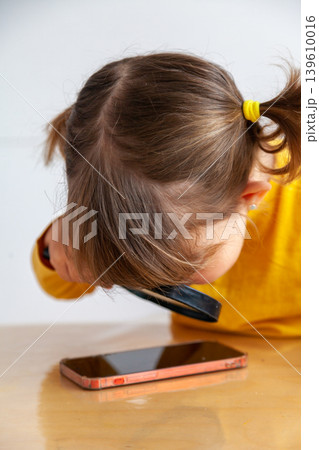A little girl examines a mobile phone with a magnifying glass, introducing concepts such as early exposure to mobile phones or internet safety. 139610016