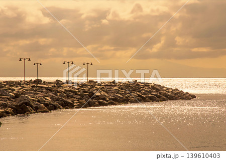 Golden Hour Coastal Breakwater with Lamps.A serene sunset view of a rocky sea wall and vintage street lights reflecting on calm ocean waters. Golden Hour Coastal Breakwater with Lamps.A serene sunset view of a rocky sea wall and vintage street lights reflecting on calm ocean waters. 139610403