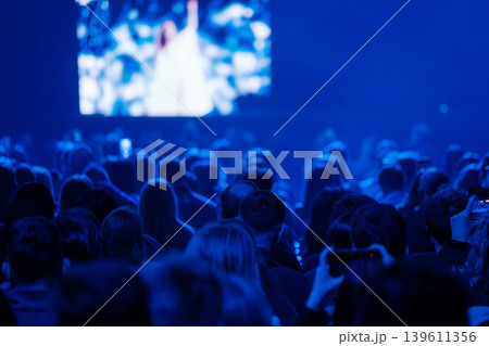 Audience watching live concert on large screen, blue stage lighting creates atmospheric mood 139611356