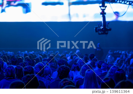 Audience watching conference presentation on large screen, stage lighting and camera rig in blue Audience watching conference presentation on large screen, stage lighting and camera rig in blue 139611364