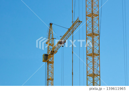 Two tall cranes stand in an urban area during the day. The cranes are positioned against a clear blue sky Two tall cranes stand in an urban area during the day. The cranes are positioned against a clear blue sky 139611965