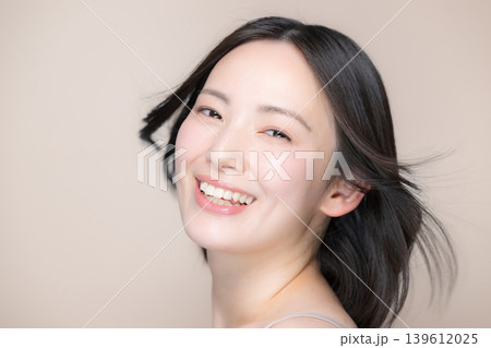 ベージュ背景の風になびくサラサラ黒髪と笑顔の女性美肌とスキンケア美容やトリートメントイメージ 139612025