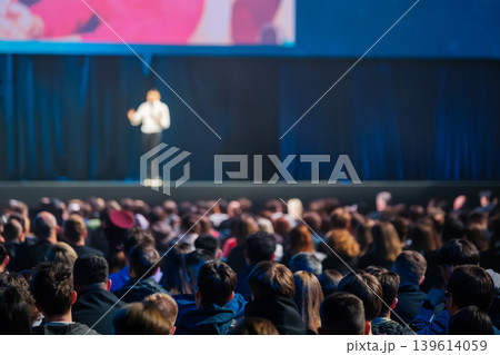 Audience watches speaker on stage at conference presentation in large dimly lit auditorium 139614059