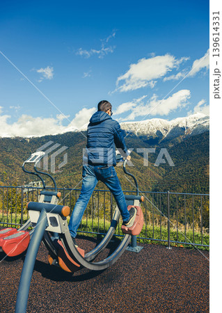Man exercises on outdoor equipment with mountain view in sunny weather Man exercises on outdoor equipment with mountain view in sunny weather 139614331