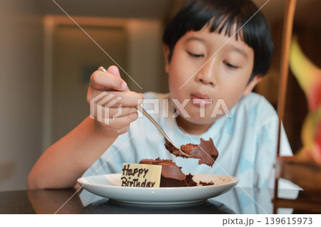 Close up of delicious chocolate birthday cake with Happy Birthday sign. Cute Asian little boy eating a piece of sweet dessert with a fork in blurred background. Joyful childhood celebration moments. 139615973