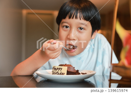 Close up of delicious chocolate birthday cake with Happy Birthday sign. Cute Asian little boy eating a piece of sweet dessert with a fork in blurred background. Joyful childhood celebration moments. Close up of delicious chocolate birthday cake with Happy Birthday sign. Cute Asian little boy eating a piece of sweet dessert with a fork in blurred background. Joyful childhood celebration moments. 139615974