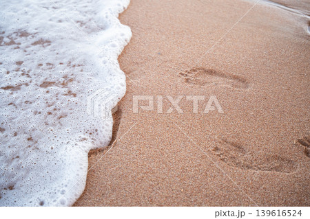 Sea wave with white foam washing over sandy beach with human footprints near the shoreline at sunset. Summer travel and coastal nature Sea wave with white foam washing over sandy beach with human footprints near the shoreline at sunset. Summer travel and coastal nature 139616524