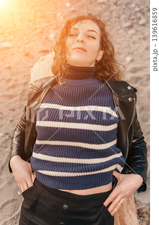 Woman beach relaxation enjoying warm golden sunlight on a pebble shore wearing a leather jacket and striped sweater Woman beach relaxation enjoying warm golden sunlight on a pebble shore wearing a leather jacket and striped sweater 139616859