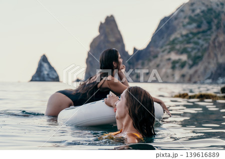 Women Inflatable Raft Beach Vacation - Two women relax on an inflatable raft in the ocean, enjoying a beach vacation with stunning mountain views. Women Inflatable Raft Beach Vacation - Two women relax on an inflatable raft in the ocean, enjoying a beach vacation with stunning mountain views. 139616889