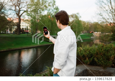 A man holds up his smartphone in a park to find a signal. The screen displays a "No Signal" icon, illustrating a cellular network outage or poor connection. A man holds up his smartphone in a park to find a signal. The screen displays a "No Signal" icon, illustrating a cellular network outage or poor connection. 139617828