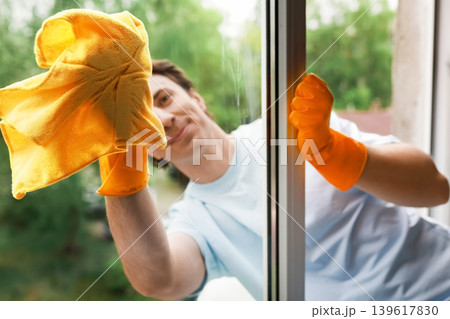 A man in orange gloves smiles while wiping a window with a yellow cloth. This bright image shows spring cleaning and home maintenance A man in orange gloves smiles while wiping a window with a yellow cloth. This bright image shows spring cleaning and home maintenance 139617830