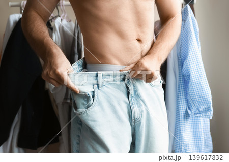 Man showing significant weight loss by pulling on his oversized jeans. Concept of body transformation, healthy lifestyle, and successful fitness progress. 139617832