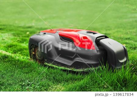 A red and black robotic lawn mower automatically trims grass on a lush green lawn. A perfect example of smart gardening and eco-friendly technology. A red and black robotic lawn mower automatically trims grass on a lush green lawn. A perfect example of smart gardening and eco-friendly technology. 139617834