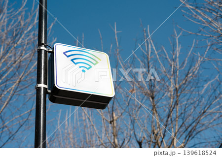 A white square sign with a colorful Wi-Fi symbol mounted on a black metal pole outdoors. The sign indicates a public wireless internet hotspot. 139618524