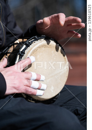 Close-up of a musician playing a djembe drum outdoors. Hands of a drummer with taped fingers playing an ethnic percussion instrument. Close-up of a musician playing a djembe drum outdoors. Hands of a drummer with taped fingers playing an ethnic percussion instrument. 139618525