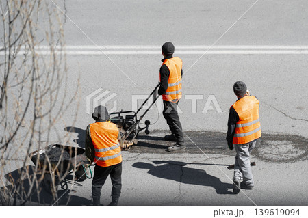Road workers in orange reflective vests repairing a pothole on a city street using a plate compactor and a wheelbarrow with fresh asphalt. A team of municipal workers performing road maintenance and Road workers in orange reflective vests repairing a pothole on a city street using a plate compactor and a wheelbarrow with fresh asphalt. A team of municipal workers performing road maintenance and 139619094