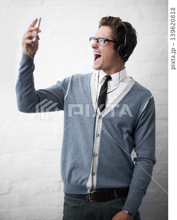 Man, headphones and singing to music and phone for rock song by wall background, voice and sound. Male person, smartphone and streaming radio or podcast, playlist and hearing audio and mobile app 139620818