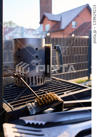 Smoking charcoal chimney starter stands on grill grate with barbecue brush and fork Smoking charcoal chimney starter stands on grill grate with barbecue brush and fork 139624213