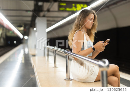 Young girl sitting in subway platform holding phone and wireless headphones on airs Young girl sitting in subway platform holding phone and wireless headphones on airs 139625633